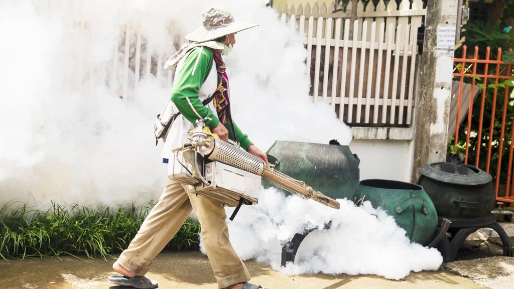 A man wearing a hat and scarf walks along a paved path next to a fence and plants, operating a large thermal fogger machine that is emitting a thick cloud of white smoke. This is a common method for outdoor mosquito control.