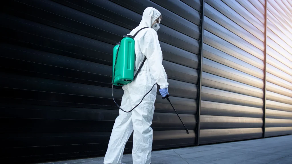 A technician from a pest control service provider in a protective suit is kneeling and spraying a chemical along an indoor wall, performing a pest control service. This is a common procedure for pest control services Malaysia.